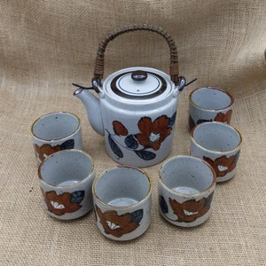 May include: A ceramic tea set featuring a teapot and six matching cups. The teapot has a woven handle and a lid. The set is decorated with a floral pattern in shades of orange, brown, and blue against a speckled, off-white background.