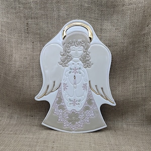 Puede incluir: Figura de ángel de cerámica con halo y detalles dorados. El ángel tiene los ojos cerrados y sostiene una vela. El vestido del ángel presenta un diseño floral en rosa y verde. Las alas son blancas con ribetes dorados.