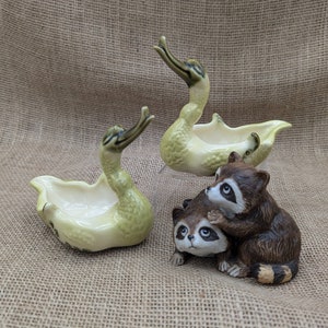 May include: Three ceramic ornaments: two swan-shaped dishes in pale green and a brown raccoon pair. The swans have raised necks and open bowls. The raccoons are in a playful embrace, with detailed fur.