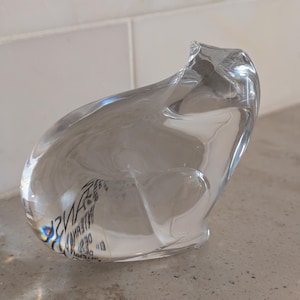 May include: A clear glass sculpture in the shape of a stylized cat. The transparent artwork has smooth curves and a slightly rough edge at the top. The sculpture is sitting on a speckled grey surface.