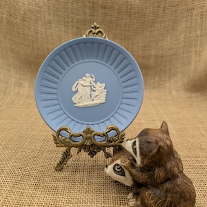 May include: A light blue decorative plate with a white relief design, displayed on a gold-colored metal stand. A brown and white raccoon figurine is positioned to the right of the plate. The plate has a ridged border.
