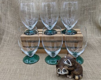 May include: Six clear wine glasses with green bases are arranged on a wooden rack. A small raccoon figurine is in the foreground. The glasses have a classic shape, suitable for serving wine. The background is a burlap fabric.