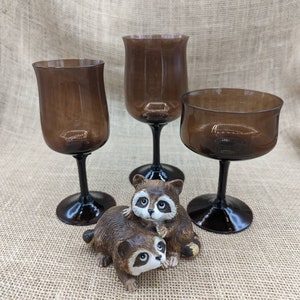 May include: Three brown-tinted glass goblets with black stems and bases are displayed alongside a raccoon figurine. The goblets vary in shape, with one being a champagne-style glass. The raccoons are brown and white.