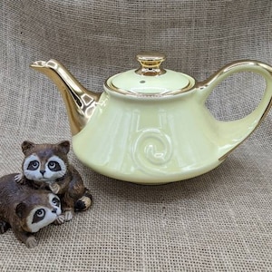Könnte beinhalten: Eine hellgelbe Teekanne mit goldenen Verzierungen und einem passenden Deckel. Die Teekanne hat ein dekoratives Wirbeldesign. Zwei kleine Waschbärfiguren befinden sich vor der Teekanne. Die Figuren sind braun und weiß mit schwarzen Augen.