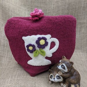 May include: A heart-shaped, burgundy tea cozy with a white teacup applique featuring purple flowers and green leaves. A small, pink crocheted flower adorns the top. Two raccoon figurines are positioned in front of the cozy.