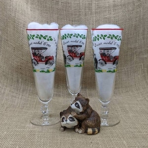 Puede incluir: Tres copas altas y transparentes con bordes rojos y diseños de coches antiguos, llenas de espuma blanca. Una figura de cerámica de mapache está delante de las copas. Las copas presentan el texto "Buick model C 1905".