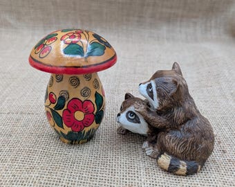 May include: A hand-painted wooden mushroom figurine with a red cap and floral designs, next to a pair of raccoon figurines. The mushroom has a red base and is decorated with red, green, and yellow flowers.