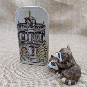 May include: A ceramic vase with a detailed illustration of a building in brown and black. Beside it, a figurine of two raccoons, one hugging the other. The vase has a light beige background.