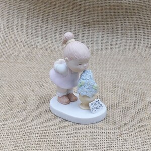 May include: A porcelain figurine of a young child in a pink dress, smelling a bouquet of light blue plastic flowers. The flowers are in a beige vase, and a sign reads "PLASTIC FLOWERS". The figurine is on a white oval base.