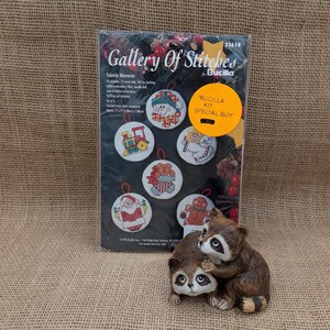 Op de afbeelding: Een Bucilla Gallery of Stitches Yuletide Memories kruissteekpakket met zes kerstornamentontwerpen. De kit bevat 14-count aida, vilt voor de achterkant, borduurgaren, naald en instructies. Een keramisch wasbeerbeeldje staat op de voorgrond.