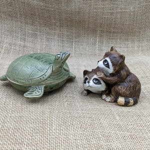 May include: A green ceramic turtle-shaped trinket box and a pair of raccoon figurines. The turtle box has a detailed shell design. The raccoons are brown with black and white markings, one on top of the other.