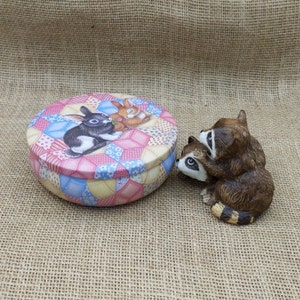 May include: A round trinket box with a patchwork design lid featuring two rabbits. Next to the box is a figurine of two raccoons. The box and figurine are on a burlap surface.