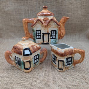 May include: A ceramic tea set featuring a teapot, sugar bowl, and creamer, all shaped like quaint cottages. The teapot has a thatched roof and a tree branch handle. The sugar bowl and creamer mirror the teapot's design, with brown and beige tones.