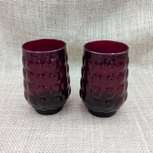May include: Two matching, deep red drinking glasses. The glasses have a textured, bubble-like pattern on the sides and a solid, dark base. The interior of the glasses is a rich, dark red. The glasses are made of glass and are suitable for beverages.