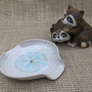 May include: A speckled, off-white ceramic spoon rest with a light blue floral design in the center. A brown raccoon figurine is in the background, with one raccoon hugging the other.