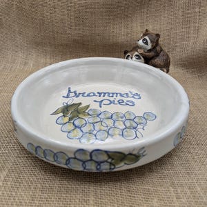 Op de afbeelding: Een witte keramische taartschaal met de woorden "Bramma's Pies" in blauw schrift, samen met een geschilderde tros druiven. De rand van de schaal heeft een herhalend druivenontwerp. Twee wasbeerbeeldjes staan op de achtergrond.