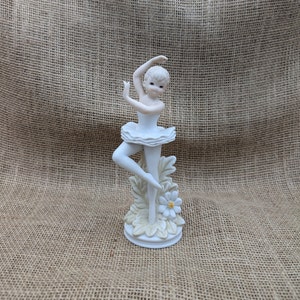 May include: A white porcelain figurine of a ballerina in a graceful pose. The ballerina wears a tutu and is posed on a base with floral and leaf accents. The figurine has a delicate, hand-painted appearance, with subtle color variations.