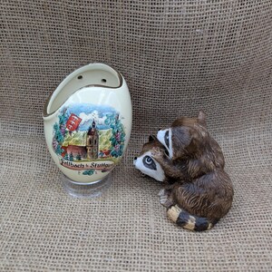 May include: A cream-colored ceramic wall pocket with a painted scene of a European town, and a small raccoon figurine. The wall pocket has a gold rim and the town scene includes a church and the text "Fellbach b. Stuttgart". The raccoon figurine is brown and white.