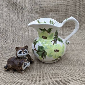 May include: A white ceramic jug with a floral design in shades of green and yellow, and a brown and white raccoon figurine. The jug has a curved handle and a wide spout. The raccoon figurine depicts two raccoons.