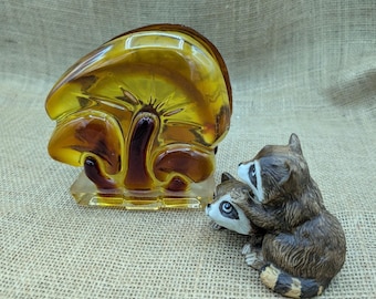 May include: A glass mushroom-shaped object with amber and brown tones, and a small figurine of two raccoons. The mushroom is on a clear base. The raccoons are brown, white, and black.