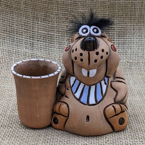 May include: A whimsical, brown ceramic figurine of a cartoon-like creature with a black tuft of hair and large eyes. It has a matching brown cup next to it. The creature has a blue and white striped collar and red accents.
