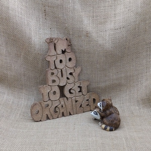 Pode incluir: Uma placa decorativa castanha com o texto "I'M TOO BUSY TO GET ORGANIZED" e uma pequena estatueta de dois guaxinins. Os guaxinins são castanhos, brancos e pretos. A placa e as estatuetas estão sobre um fundo de serapilheira.