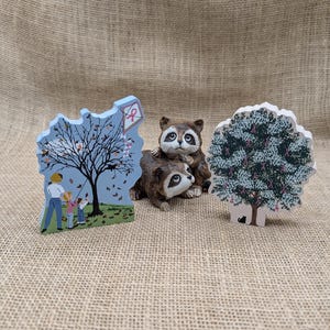May include: Three decorative wooden figurines: a blue scene with a family flying a kite, two raccoons, and a tree with pink ribbons. The figurines are set against a neutral burlap background.
