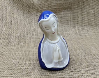 May include: A ceramic figurine of a religious figure with a blue head covering and white veil. The figure has closed eyes and clasped hands, with a gold-colored necklace. The figurine is on a neutral-colored textured surface.