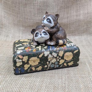 May include: A decorative box with a floral design and two raccoon figurines. The box is rectangular with a dark green background and cream-colored flowers. The raccoons are brown and white.