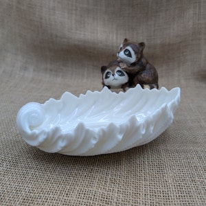 May include: A white, leaf-shaped dish with a ruffled edge, and a figurine of two raccoons. The raccoons are brown and white, with one perched atop the other. The dish is made of a glossy, opaque material.
