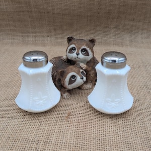 May include: Two white glass salt shakers with silver lids, decorated with floral patterns, flank a ceramic raccoon figurine. The figurine features two raccoons, one hugging the other. The background is a textured, tan fabric.