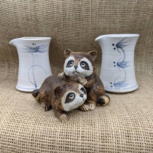 May include: A still life featuring two white ceramic pitchers with blue floral designs and two raccoon figurines. The raccoons are brown and tan with black markings. The pitchers have a curved spout.