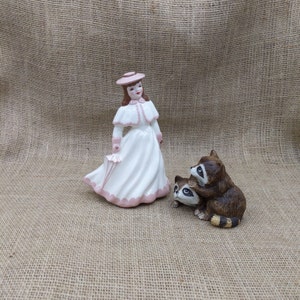 Puede incluir: Figura de cerámica de una mujer con vestido blanco y detalles rosas, sosteniendo un paraguas rosa. Junto a ella, dos mapaches marrones. Las figuras están sobre una superficie de arpillera beige.