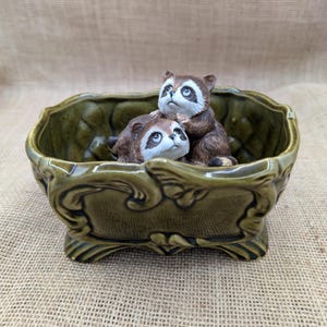 May include: A decorative ceramic planter in a muted green colour, featuring a rectangular shape with ornate detailing. Two raccoon figurines with brown fur and white faces are nestled inside, creating a charming, whimsical scene. The planter is elevated on small feet.
