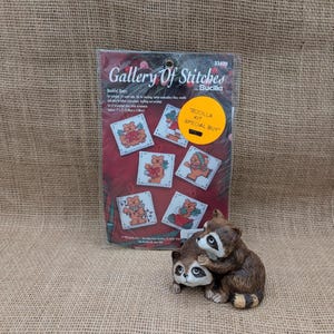 May include: A Bucilla "Gallery of Stitches" counted cross-stitch kit featuring six square designs with teddy bears. A ceramic raccoon figurine is in front of the kit. The kit has a "Special Buy" sticker.