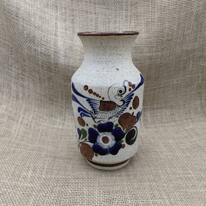Puede incluir: Un jarrón de cerámica con una base moteada de color blanquecino y un borde marrón. El jarrón está decorado con motivos florales y de pájaros azules, junto con detalles marrones y verdes. El jarrón tiene una parte superior ligeramente acampanada y un cuerpo redondeado.