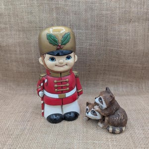 May include: A ceramic figurine of a soldier wearing a gold helmet with green holly leaves and a red uniform with white trim, holding a candy cane, positioned next to a smaller ceramic figurine of two brown and white raccoons sitting together.