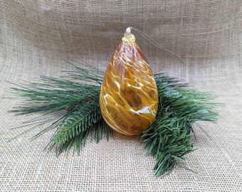 May include: A teardrop-shaped Christmas ornament with a brown and white marbled design. The glass ornament hangs from a silver string and rests on a bed of green pine needles. The background is a tan burlap.