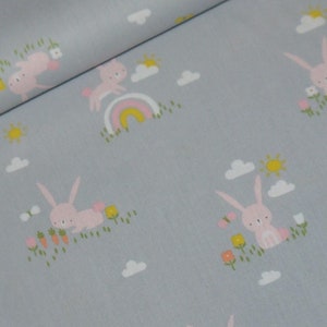 May include: A light blue fabric with a repeating pattern of pink bunnies hopping over a rainbow, clouds, and flowers. The bunnies have white ears and tails.