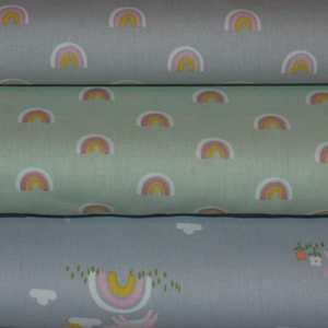 May include: Four pieces of fabric with different patterns. The top fabric is light blue with a white and pink rainbow pattern. The second fabric is light green with a white and pink rainbow pattern. The third fabric is light blue with a white cloud and sun pattern and pink bunnies. The bottom fabric is light blue with a white cloud and sun pattern and pink bunnies.