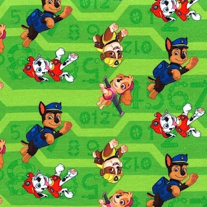 Könnte beinhalten: Ein grüner Stoff mit einem sich wiederholenden Muster aus Cartoon-Hunden aus der Fernsehsendung Paw Patrol. Die Hunde tragen ihre charakteristischen Uniformen und sind von Zahlen umgeben.