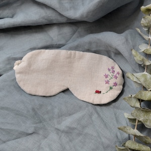 May include: A beige linen sleep mask with a small embroidered flower design and a red ladybug. The mask is on a light blue fabric background.