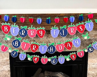 May include: A colorful banner with the words "Berry First Birthday" in red, blue, and purple letters. The banner is decorated with strawberries, blueberries, and leaves. The numbers 1 through 10 are also included on the banner.