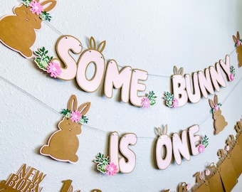 Puede incluir: Una pancarta rosa y marrón con el texto "Some Bunny Is One" y recortes de conejos. La pancarta está decorada con flores rosas y hojas verdes.
