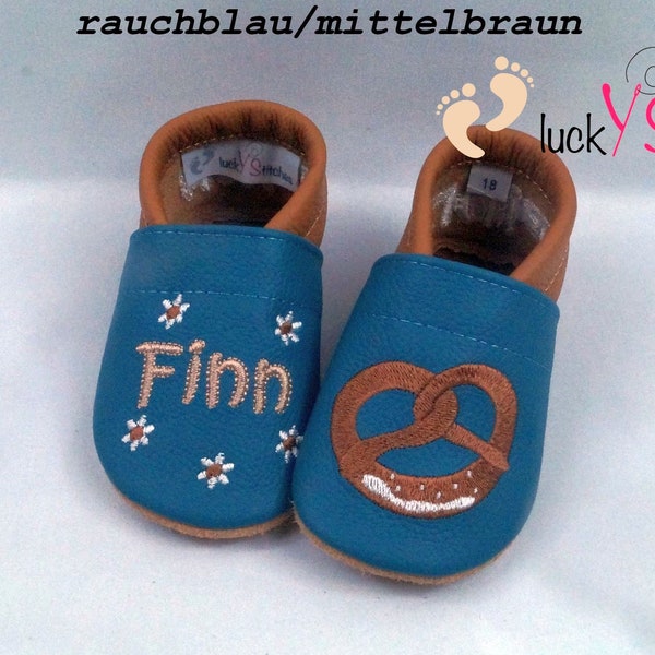 May include: A pair of teal blue baby shoes with brown leather trim. One shoe has the name "Finn" embroidered on it, and the other has a brown pretzel embroidered on it. The shoes are decorated with white daisy flowers.