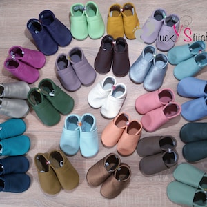Könnte beinhalten: Eine Sammlung von 24 Paar weichen Lederschühchen für Babys in verschiedenen Farben, darunter Grün, Blau, Braun, Rosa und Weiß. Die Schuhe sind in einem Kreis auf einer Holzoberfläche angeordnet. Der Text "luckystitches" ist in der oberen rechten Ecke des Bildes sichtbar.