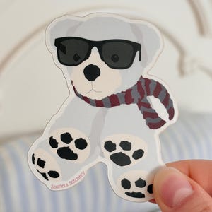 Könnte beinhalten: Ein Aufkleber mit einem Cartoon-Eisbären, der eine schwarze Sonnenbrille und einen rot-grau-weiß gestreiften Schal trägt. Der Bär hat schwarze Pfotenabdrücke auf seinen Füßen. Der Text "Scarlet's Stitchery" ist aufgedruckt.