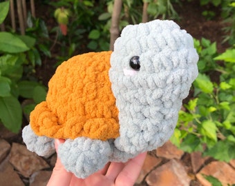 May include: A crocheted turtle toy with a gray shell and orange underbelly. The toy has a single black eye and a stitched smile.
