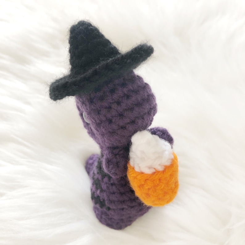 May include: A handmade crochet witch doll in purple with a black hat. The doll is holding an orange and white candy corn basket. The witch has black stripes on its body, perfect for Halloween decorations.