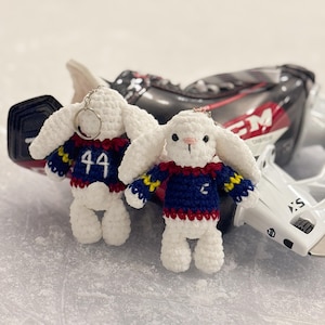 May include: Two white crocheted rabbit keychains with long ears, each wearing a dark blue sweater with red and yellow trim. One rabbit shows the number 44 on the back of its sweater, while the other displays a small white letter C on the front.